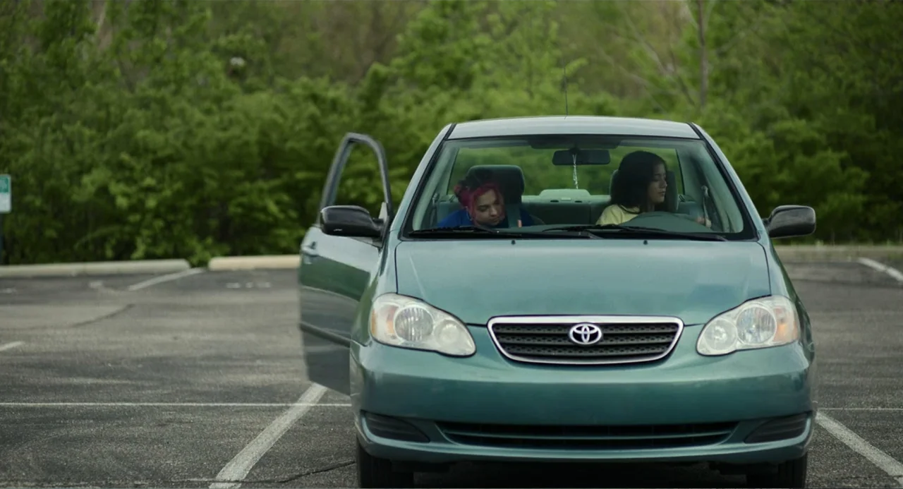 Turtles All the Way Down (2024) movie still: medium — A teal sedan sits in an asphalt parking lot backed by dense green trees. Two young women are…