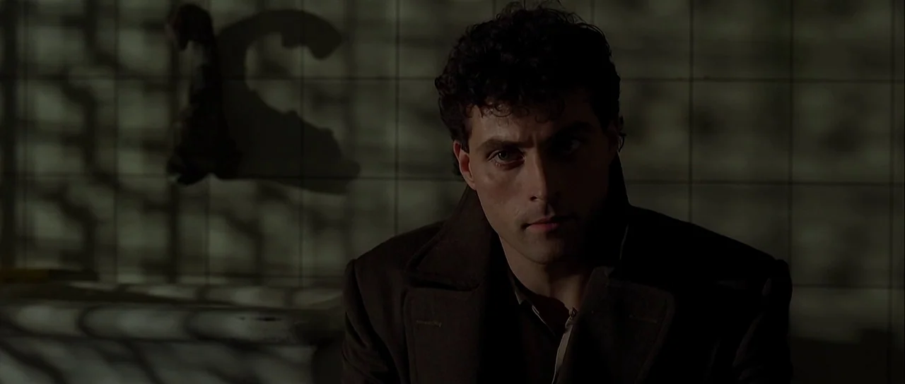 Dark City (1998) movie still: medium — A tiled room features a dark, patterned shadow cast across the wall and subject. A man in a dark coat sits…