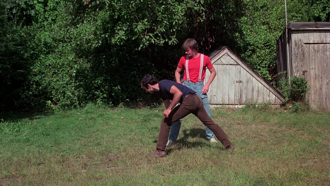 I Spit On Your Grave (1978) movie still: fullbody — A grassy backyard features a small wooden storm cellar door and dense trees. One man stands…
