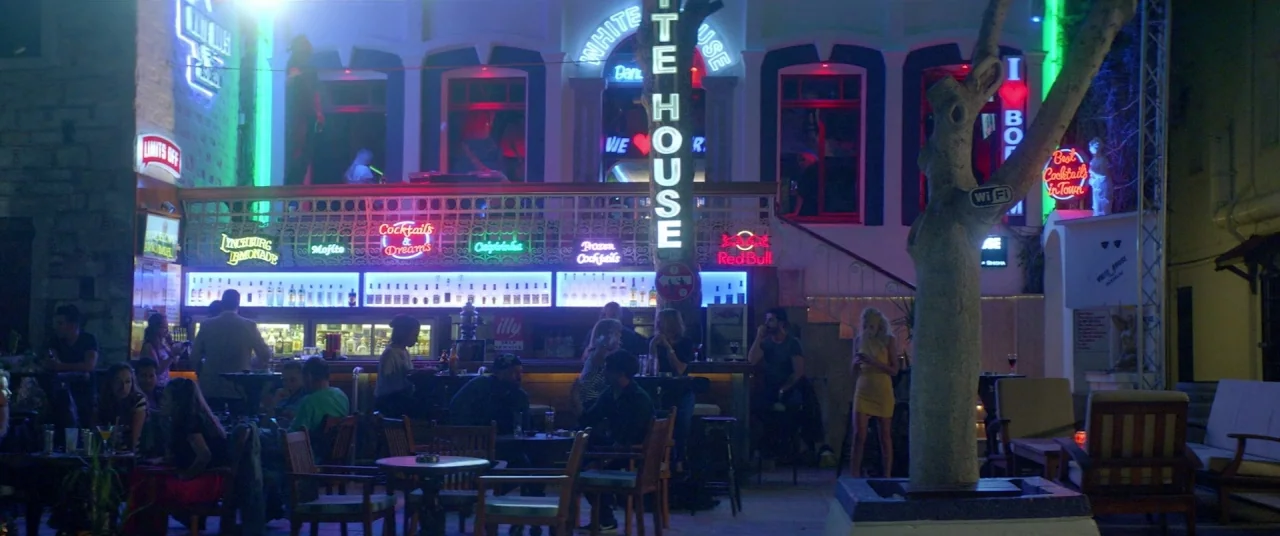 Holiday movie still: wide — A multi-level bar exterior at night features neon signage and an outdoor seating area. People sit at small tables on the…