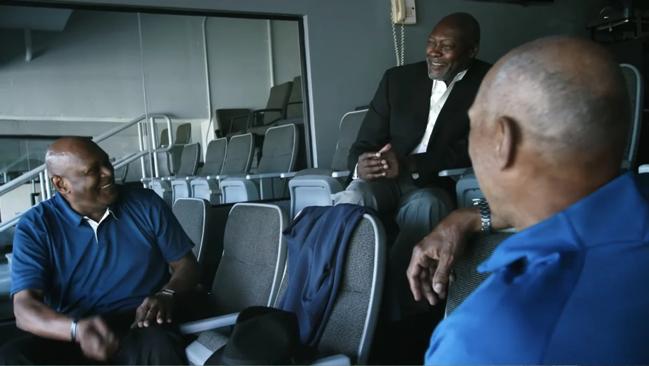 Reggie (2023) movie still: medium — A luxury stadium suite features rows of gray upholstered seats and a large glass wall. Three older men sit in…