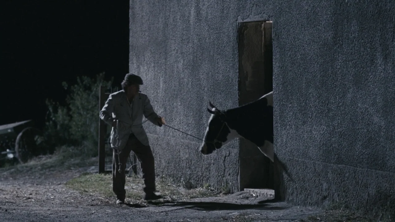 Shadows in the Sun (2005) movie still: fullbody — A man stands outside a textured concrete building at night on a dirt path. He holds a rope that is…