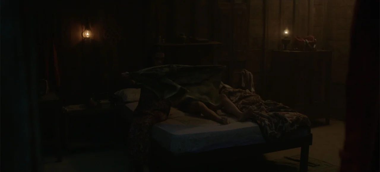 A Thousand Days (2023) movie still: medium — A dimly lit room features wooden walls and an unmade bed with patterned blankets. A woman sits on the…