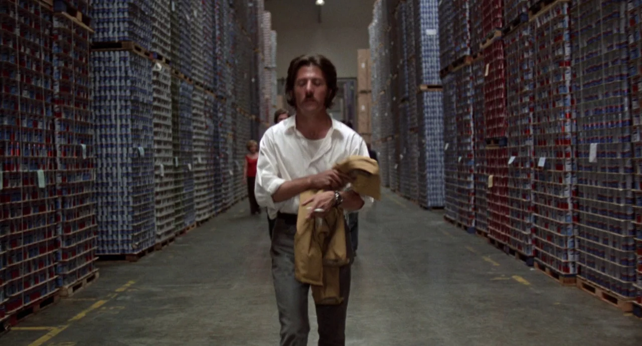 Straight Time (1978) movie still: fullbody — A vast industrial space is filled with towering stacks of inventory on wooden pallets. A man with a…