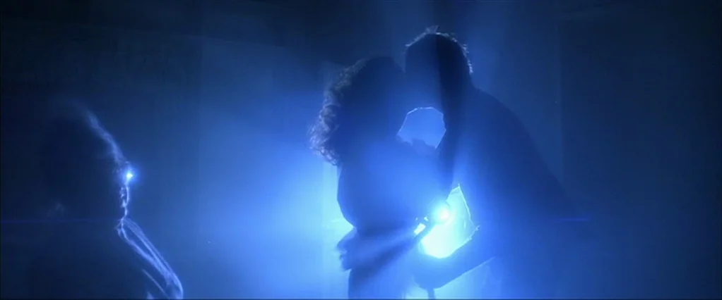 Poltergeist movie still: medium — A dark indoor setting is filled with heavy blue light and visible haze. A couple kisses in the center while an…