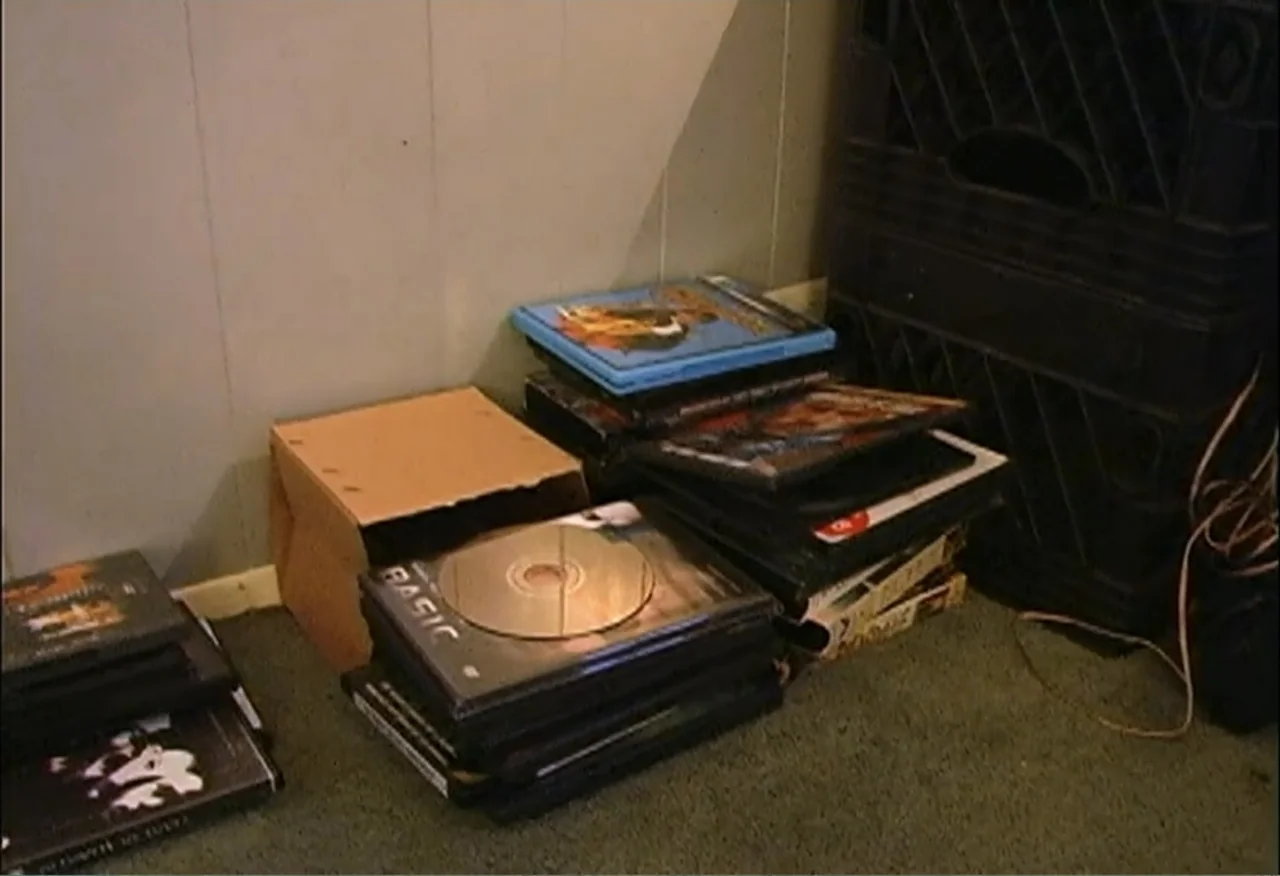Dope Sick Love (2005) movie still: medium — A pile of various DVD cases and a small cardboard box rest on carpet. The items are stacked against a…