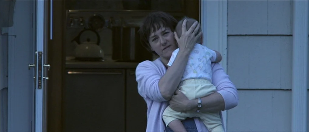 Session 9 (2001) movie still: medium — A woman stands outside a house at dusk holding a small infant. The background shows a dimly lit interior with…