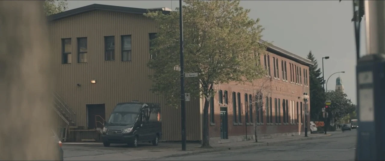 Zoe (2018) movie still: establishing — A city street corner shows a corrugated metal building and a brick structure. A black van is parked near the…