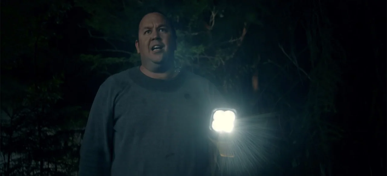 Boar (2018) movie still: medium — A dense patch of dark woods at night filled with blurred tree branches. A middle-aged man stands in the dark…