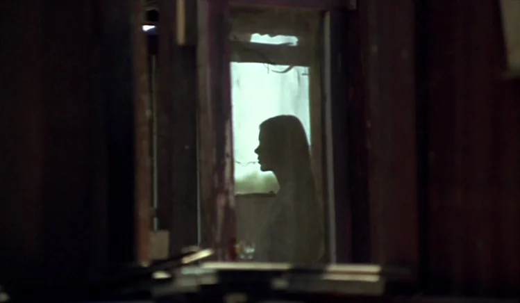 Walkabout (1971) movie still: medium — The silhouette of a young woman is visible through a dark wooden frame. She stands before a window covered by…
