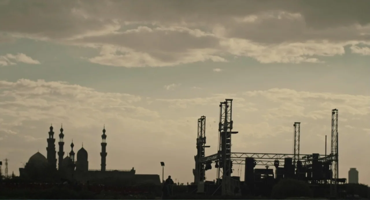 Cairo Jazzman (2017) movie still: establishing — An open field at sunset reveals the silhouettes of a mosque and stage scaffolding. The sky contains…