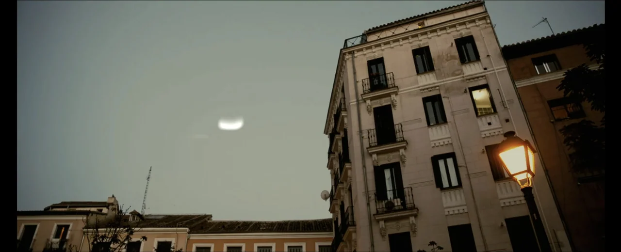 Faraday (2013) movie still: establishing — A tall European residential apartment building sits beside a shorter low-rise building at dusk. An…