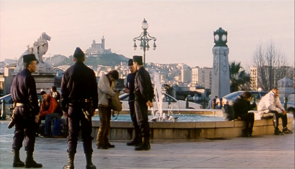 Nenette et Boni (1996) movie still: wide — A public plaza in Marseille features a circular fountain and an ornate street lamp. Police officers…