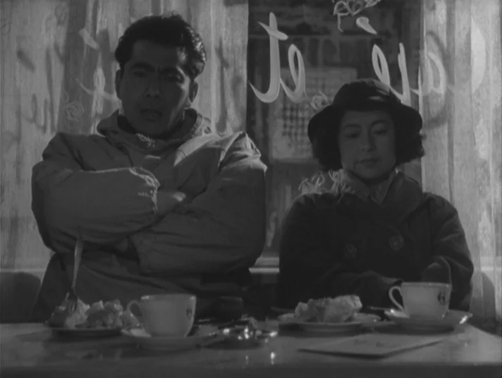 Scandal (1950) movie still: medium — A dimly lit restaurant interior viewed through a window with painted text on the glass. A man and a woman sit…