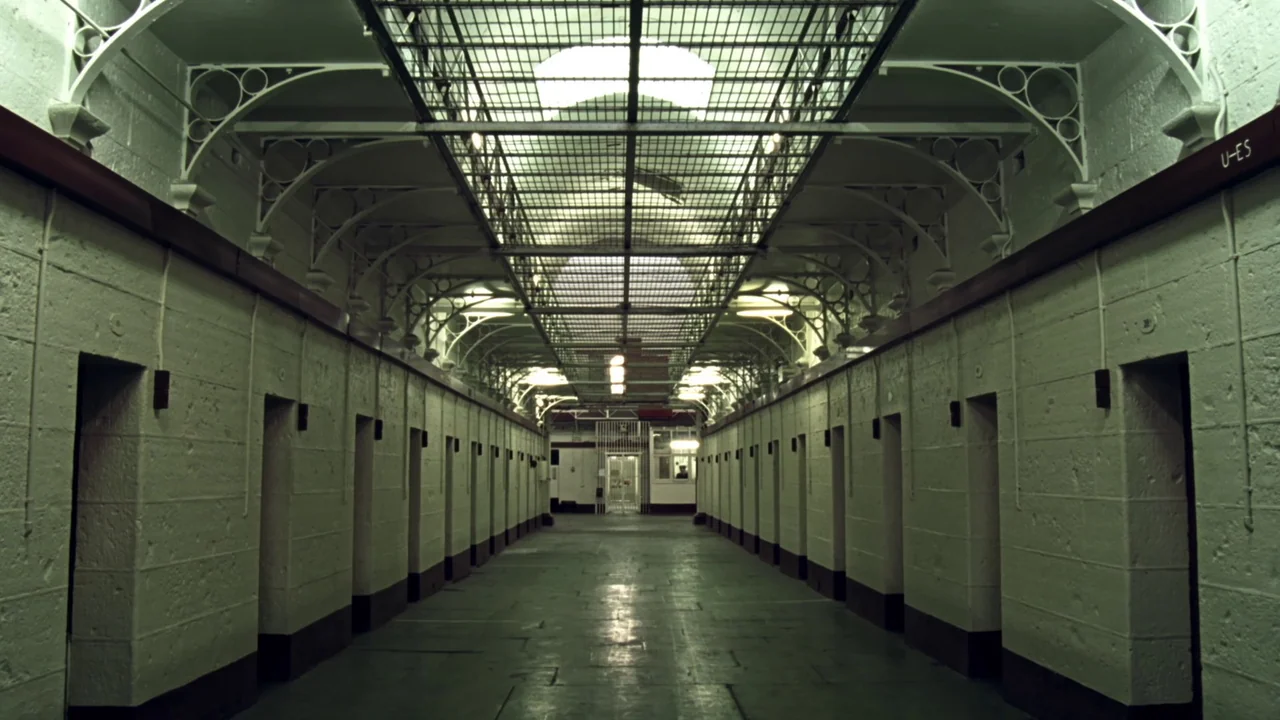Chopper (2000) movie still: establishing — A long, symmetrical prison hallway features rows of uniform doors and arched structural supports. The…