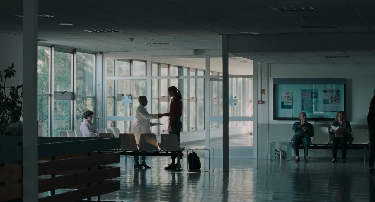 La Abuela (2021) movie still: wide — A quiet hospital waiting area with rows of plastic chairs and large floor-to-ceiling windows. A doctor and a…