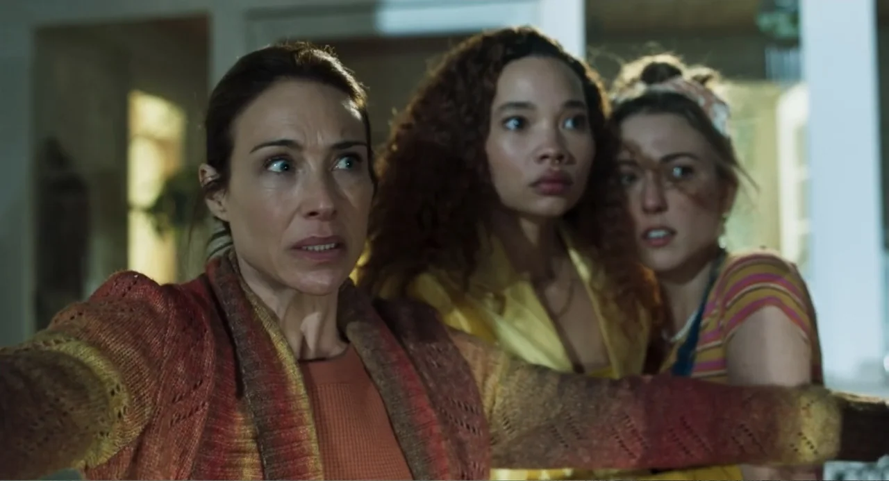 Salvation (2025) movie still: medium — A dimly lit indoor space with blurred architectural elements visible in the background. Three women stand…