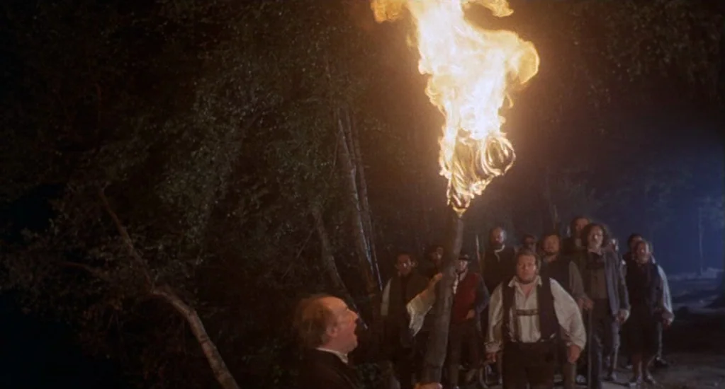 Demons of the Mind (1972) movie still: medium — A group of men moves through a dark, dense woodland at night. A leader in the foreground holds a…