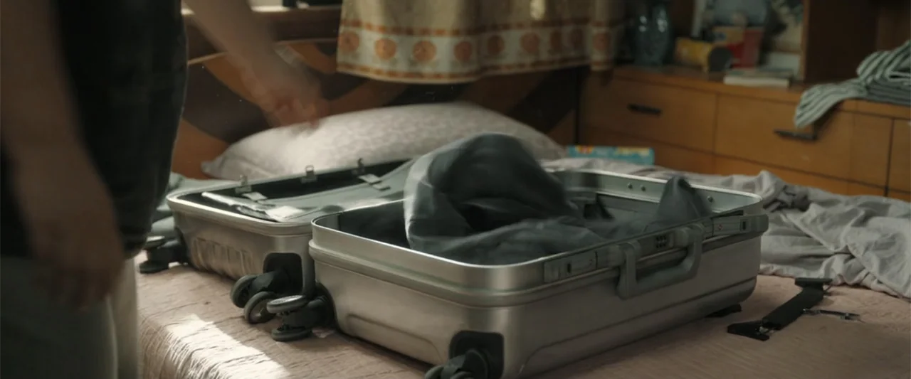 Yolo (2024) movie still: medium — A bedroom contains a bed with a rumpled sheet and an open metal suitcase. A person stands beside the bed, placing…