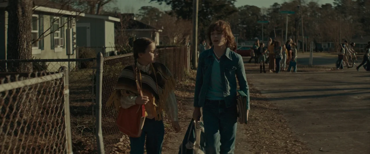 The Black Phone (2021) movie still: fullbody — A quiet residential street in a 1970s suburb features small homes behind chain-link fences. Two…