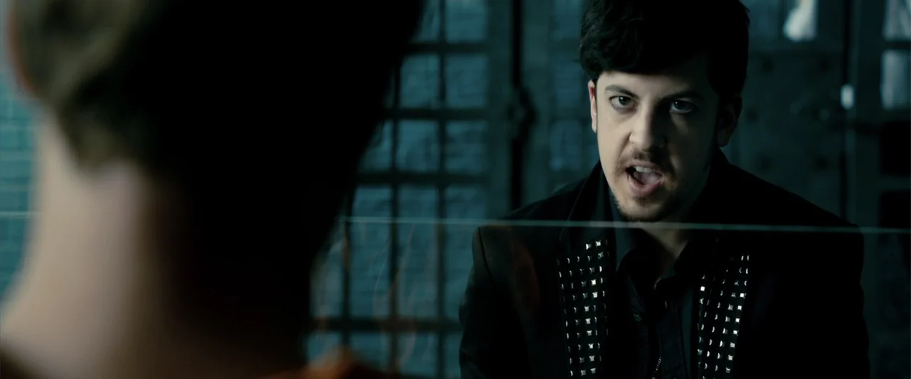 Kick-Ass 2 (2013) movie still: medium — A dark, partitioned room features a heavy metal grid behind a glass barrier. A young man with studded…