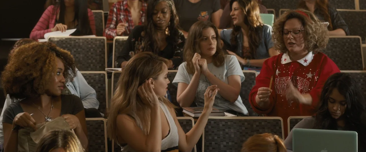 Life of the Party (2018) movie still: medium — A college lecture hall with tiered seating filled with students. A woman wearing a red sweater holds…