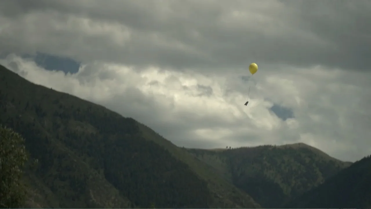 Boathouse Detectives (2010) movie still: wide — A yellow balloon ascends into a cloudy sky above dark, green-covered mountain slopes. The landscape…