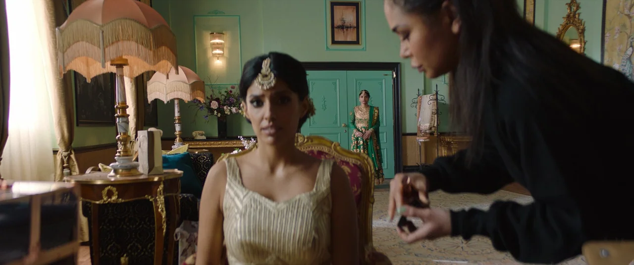 Polite Society (2023) movie still: medium — A spacious room with mint green walls features two table lamps and a standing woman. A woman sits in the…