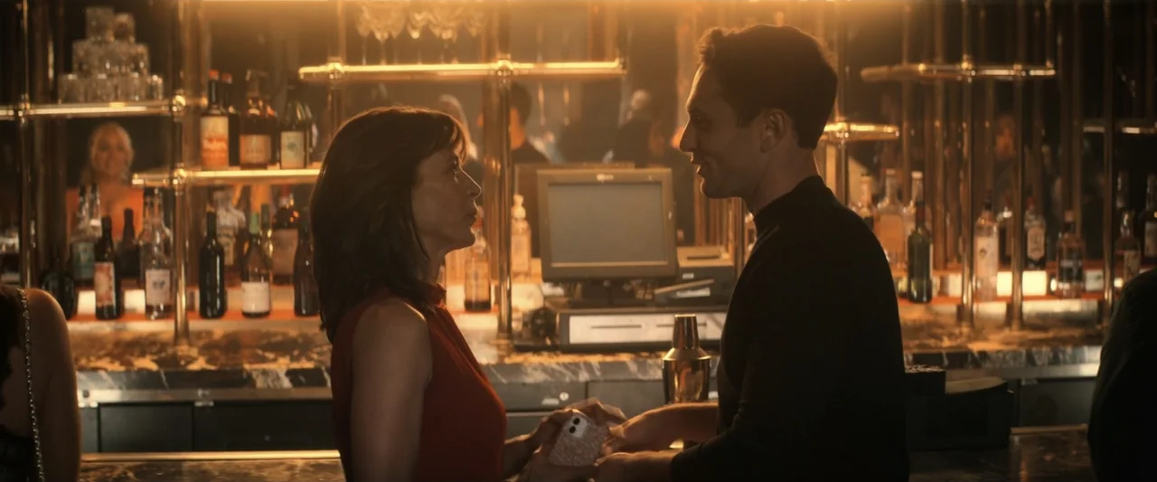 I Love America (2022) movie still: medium — A dimly lit bar features a long marble counter stocked with various glass liquor bottles. A man and a…