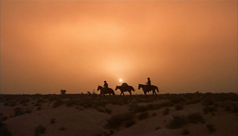 The Searchers movie still: wide — A vast, flat desert landscape under an orange sunset with sparse, dry vegetation. Three silhouettes of people on…