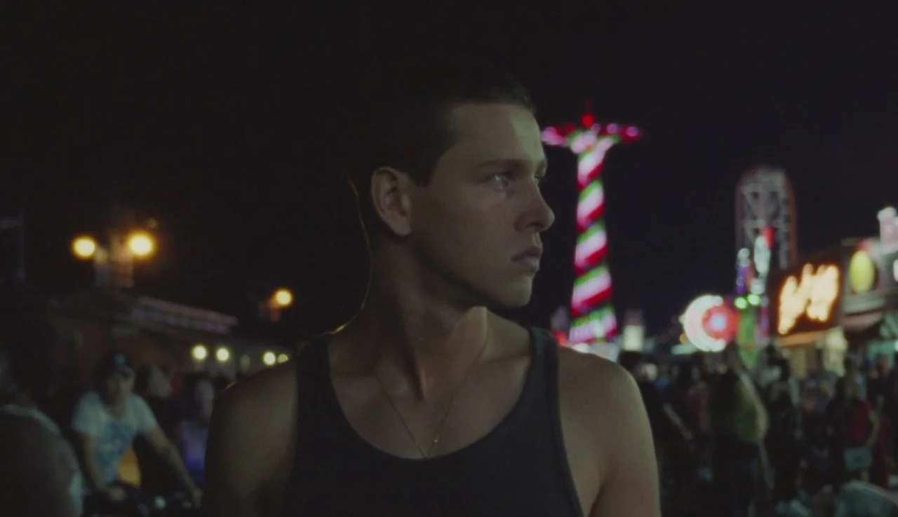 Beach Rats (2017) movie still: medium — A nighttime scene at an amusement park filled with blurred lights and a crowd. A young man wearing a tank…