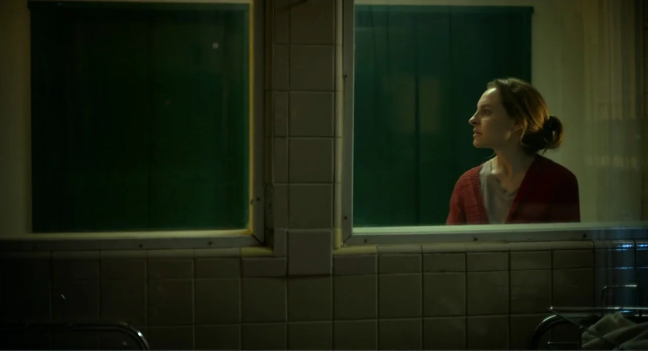 Heartbeat (2008) movie still: medium — A woman stands behind a large glass window in a room with light colored tiling. She observes the dark room on…