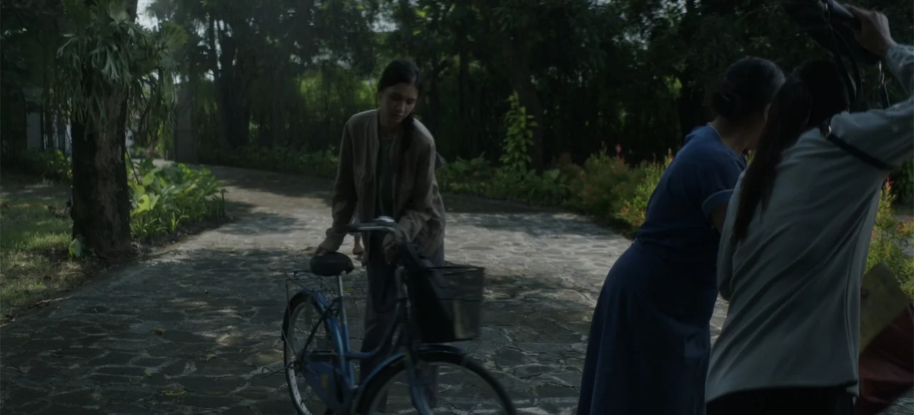 A Thousand Days (2023) movie still: fullbody — A stone-paved driveway is bordered by dense trees and green foliage in daylight. A young woman walks…