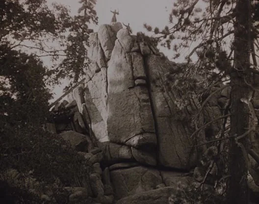 Birth of a Nation (1915) movie still: wide — A tall, jagged rock formation rises amidst a sparse pine forest. Two tiny figures stand atop the…