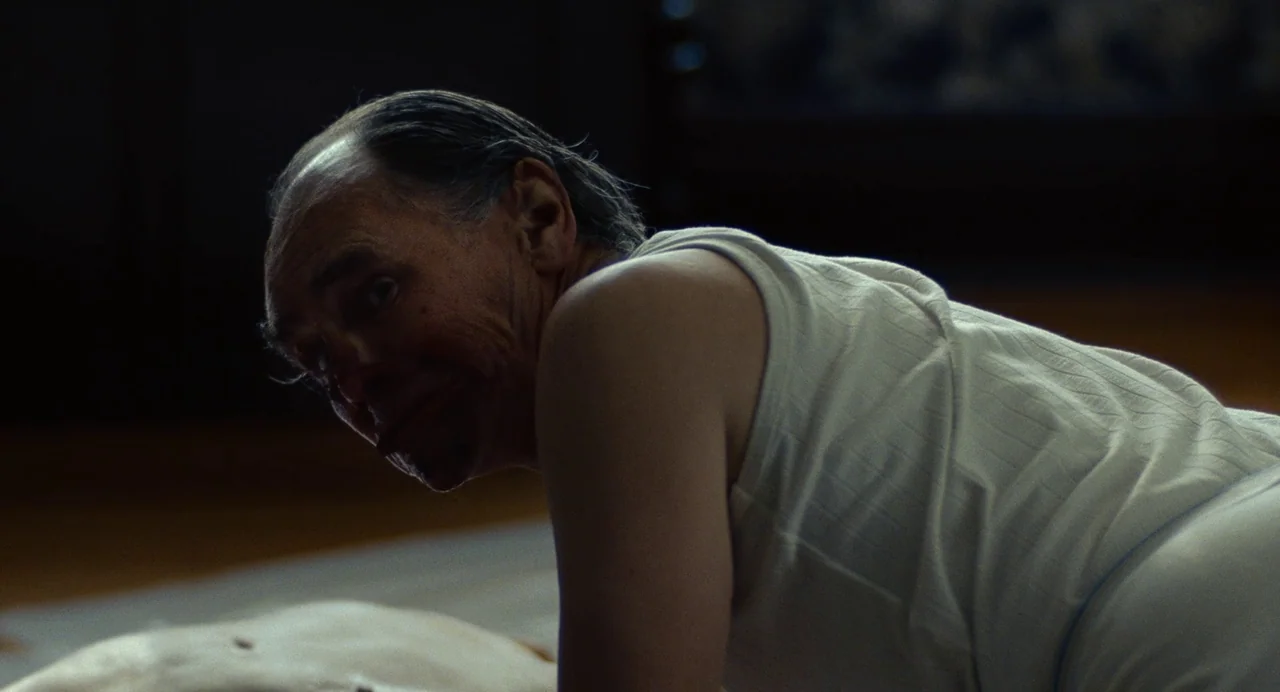 Bones and All (2022) movie still: medium — An interior space with dim, low-key lighting and a dark, indistinct background. An older man in a white…