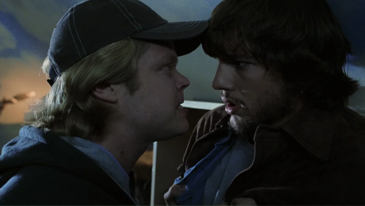 The Butterfly Effect (2004) movie still: closeup — An interior space with dim, low-key lighting focused on two men in confrontation. The men stand…