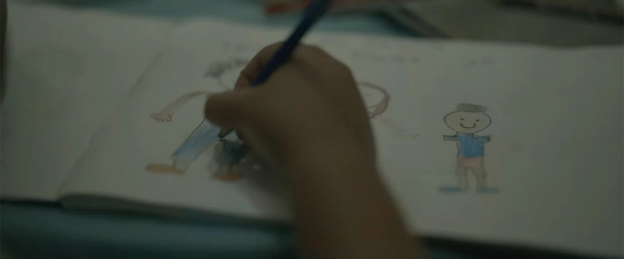 Divine Intervention (2026) movie still: closeup — A sketchbook lies open on a surface with a child's crayon drawing visible. A human hand holds a…