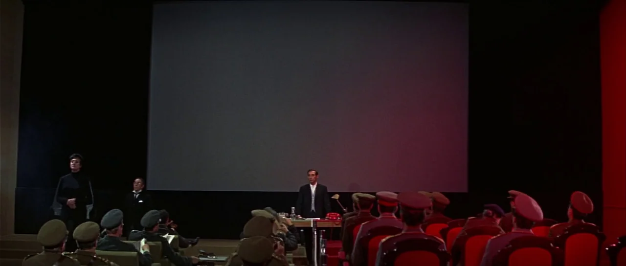 Casino Royale (1967) (1967) movie still: wide — A dark, formal briefing room features a large projection screen centered at the front. A man in a…