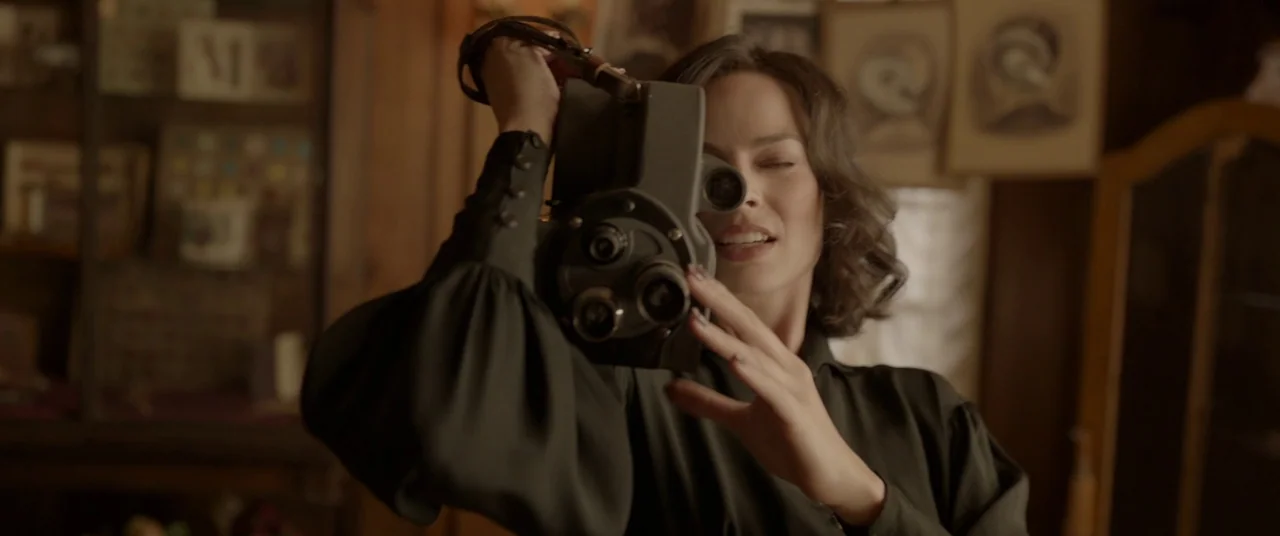 Amsterdam (2022) movie still: medium — An interior space contains vintage photographs and furniture in a softly lit room. A woman looks toward a…