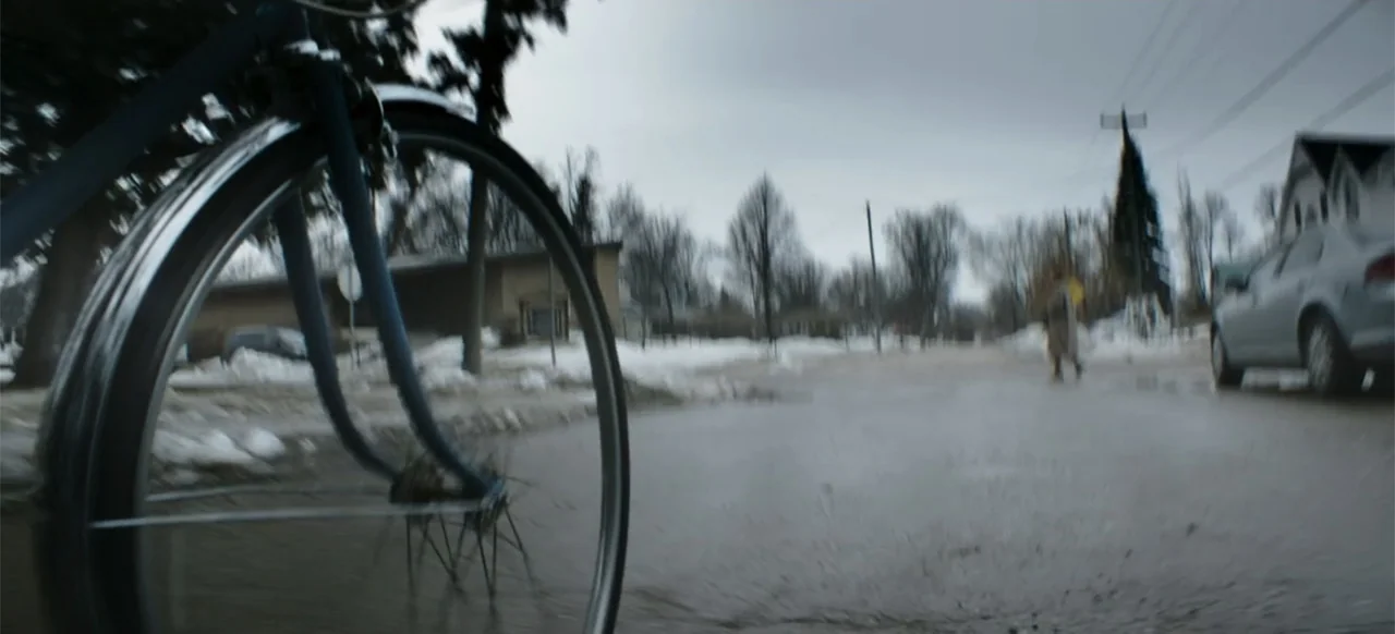 I Still See You (2018) movie still: closeup — A wet asphalt residential road with patches of melting snow on the sidewalks. A close-up view focuses…