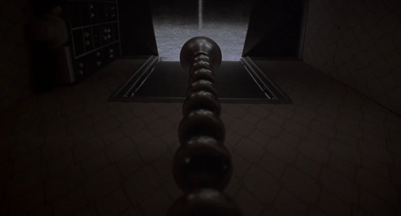 Bound (1996) movie still: closeup — A narrow dimly lit corridor features a repeating spherical pillar running down its center. The dark space is…