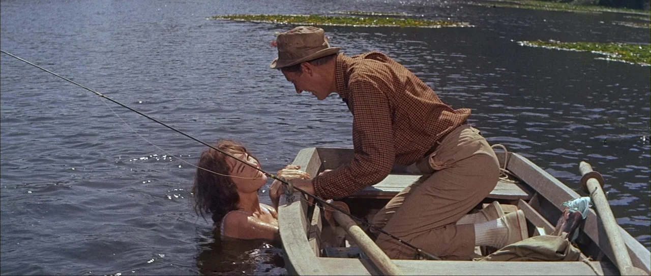 What A Way To Go! (1964) movie still: medium — A wooden rowboat floats on a lake with dark water and scattered lily pads. A man in a hat leans over…