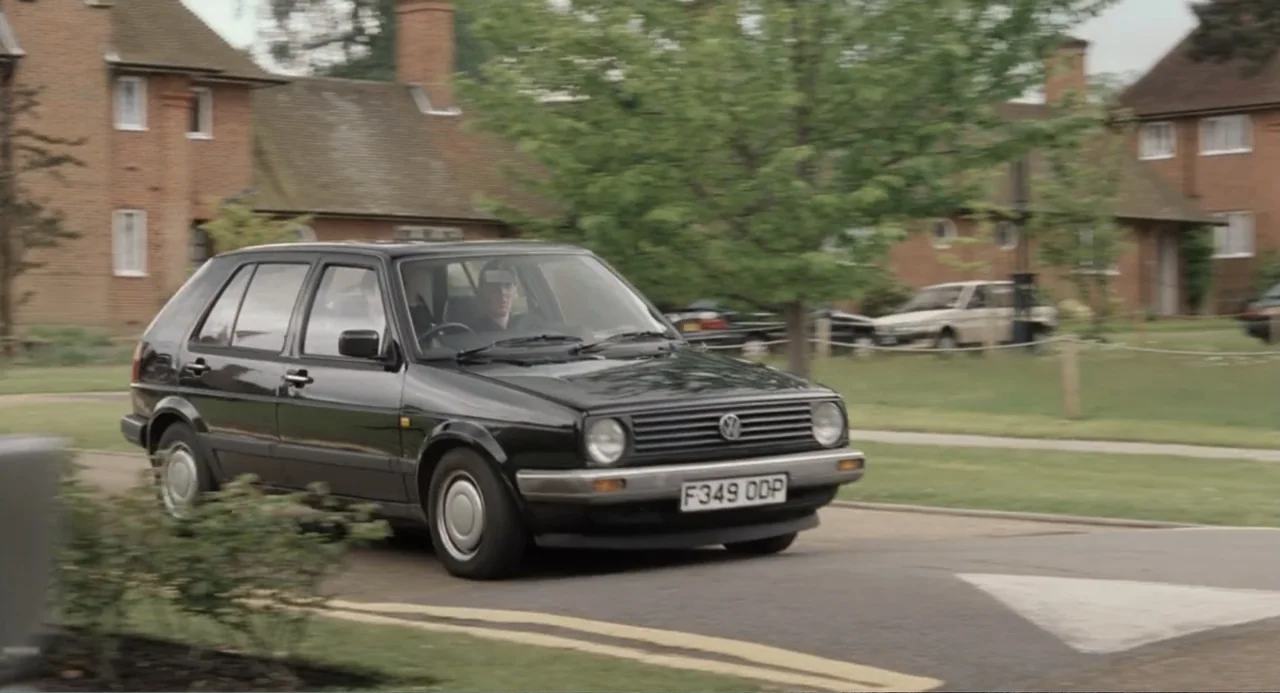 Death at a Funeral (2010) movie still: medium — A suburban street lined with brick houses and green trees under a cloudy sky. A person drives a…