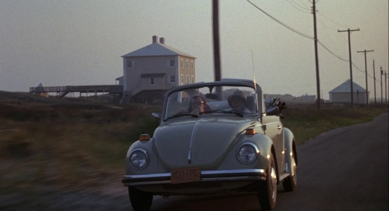 Annie Hall (1977) movie still: wide — A light-colored vintage convertible drives along a lonely stretch of coastal road at sunset. Beside the road…