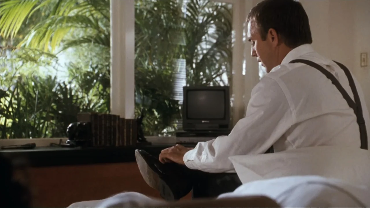 The Bodyguard (1992) movie still: medium — A sunlit bedroom features a large window overlooking dense green foliage. A man sits on the edge of the…