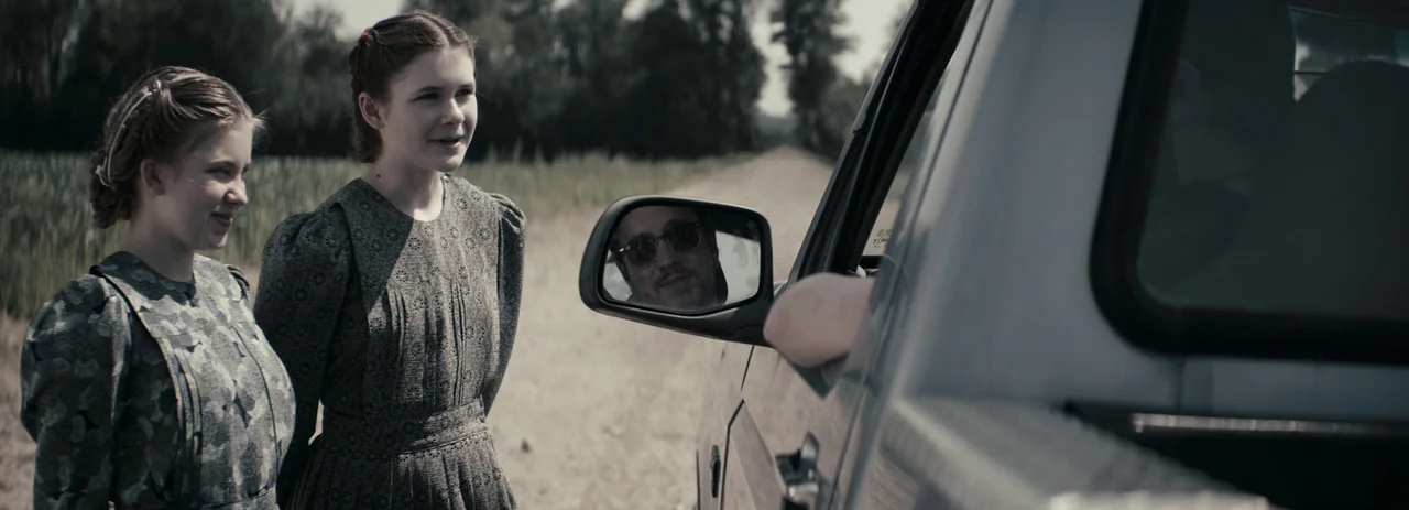 Women Talking (2022) movie still: medium — Two young women in modest floral dresses stand on a rural dirt road. A passenger in a nearby vehicle is…