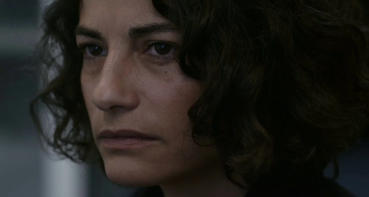 Bastards (2013) movie still: closeup — A close-up view of a woman inside a dimly lit professional space. She gazes past the camera lens with a…