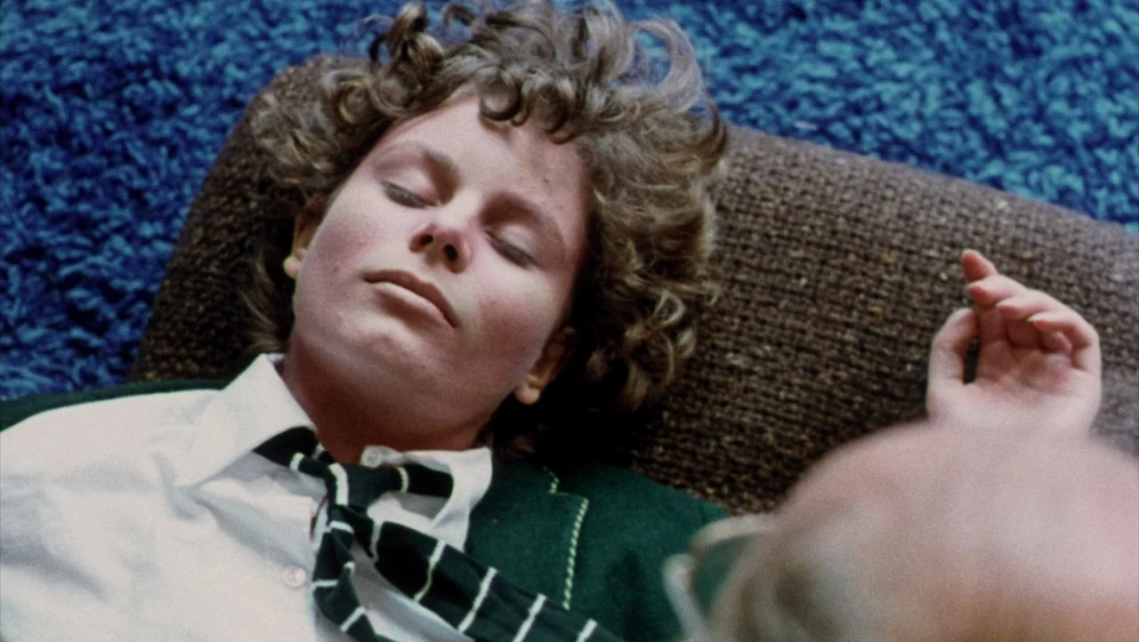 Shivers (1975) movie still: closeup — A person lies on a dark brown surface atop a thick blue carpet. The subject wears a school uniform with a…