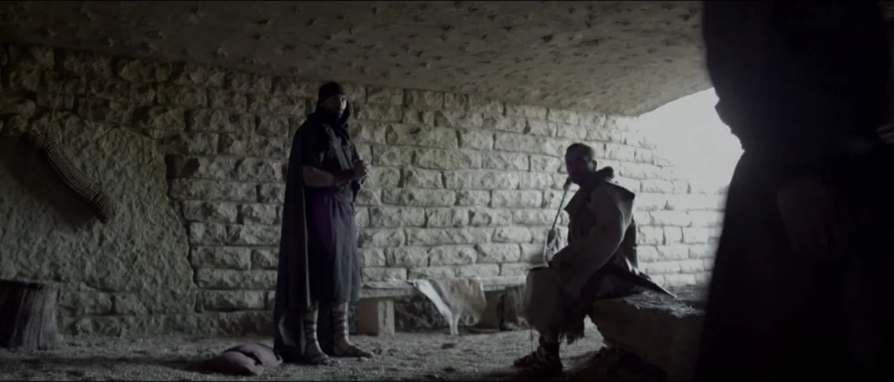 The Penitent Thief (2021) movie still: medium — A dimly lit stone chamber features rough-hewn walls and a dirt floor. Two men stand and sit inside…