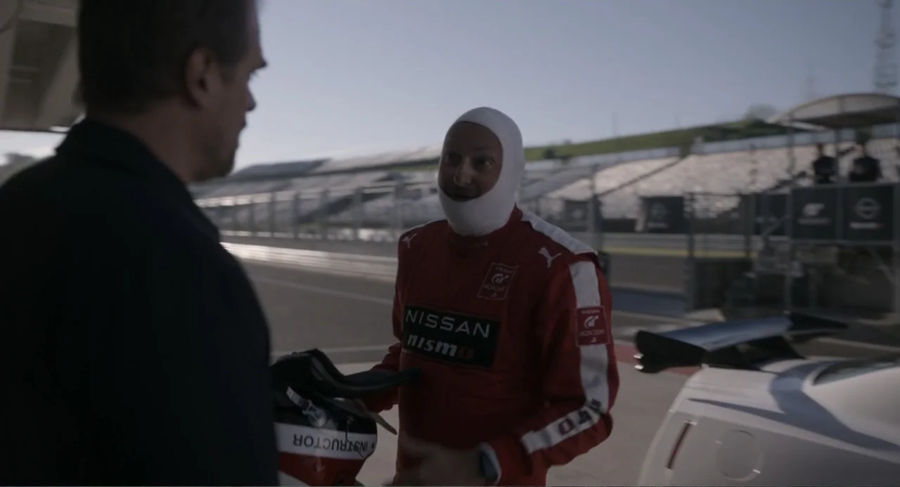 Gran Turismo (2023) movie still: medium — A sunny outdoor asphalt racing circuit features grandstands and a white car. A man in a red racing suit…